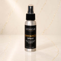 A silver spray bottle of Livingood Products Enhance Spray with a black cap and black label, standing on a light marble surface with a warm, softly lit background.