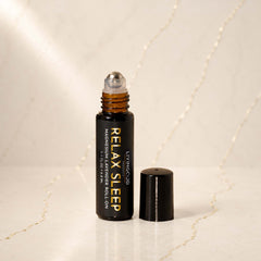 A small amber glass roller bottle with a black cap standing beside it on a light marble surface. The bottle is labeled 'LIVINGOOD RELAX SLEEP MAGNESIUM & LAVENDER ROLL ON'.