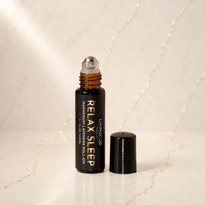 A small amber glass roller bottle with a black cap standing beside it on a light marble surface. The bottle is labeled 'LIVINGOOD RELAX SLEEP MAGNESIUM & LAVENDER ROLL ON'.