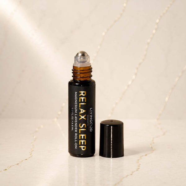 A small amber glass roller bottle with a black cap standing beside it on a light marble surface. The bottle is labeled 'LIVINGOOD RELAX SLEEP MAGNESIUM & LAVENDER ROLL ON'.